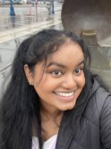 A young indian woman with long dark brown hair pulled back into a ponytail wearing a black puffer jacket smiles at the camera in front of a rainy street.